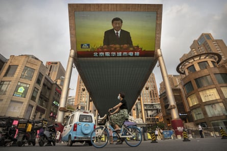 A woman wearing a face mask rides a bicycle past a large television screen at a shopping center displaying Chinese state television news coverage of Chinese President Xi Jinping