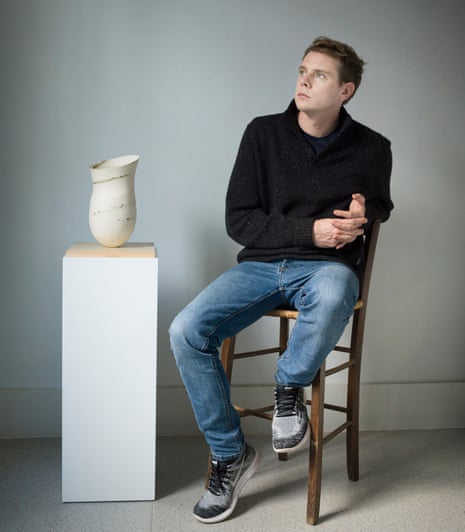 Jonathan Anderson sitting on a chair next to a plinth with a stoneware pot on top.