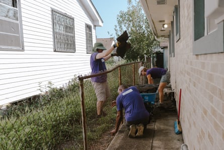 a group of three people squatting and standing doing home repairs in the exterior of a home near a grassy patch