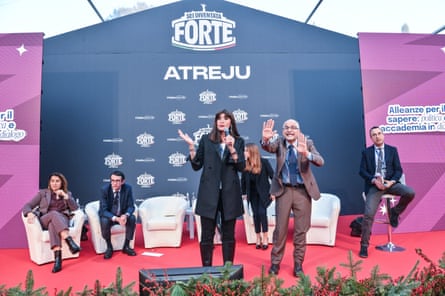 Anna Maria Bernini stands and speaks on a stage alongside a man in a brown suit; others sit in the background.