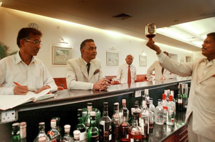 Men in white suits gather around a bar where a man holds up a bottle of alcohol