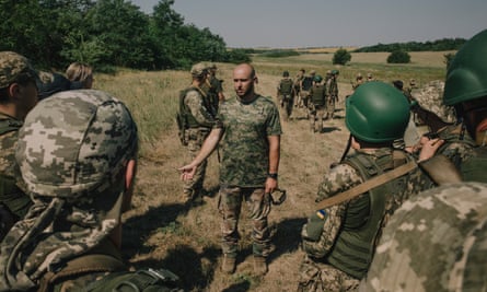 A group on recruits in camouflage clothes stand in a wide open field. Some are being given feedback by the instructor, Denis Kravchenko