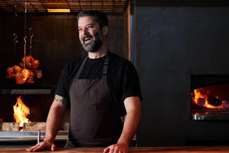 Chef Ben Williamson, dressed in a brown apron and black T-shirt, laughs in his restaurant kitchen while a wood-fire grills burns in the background