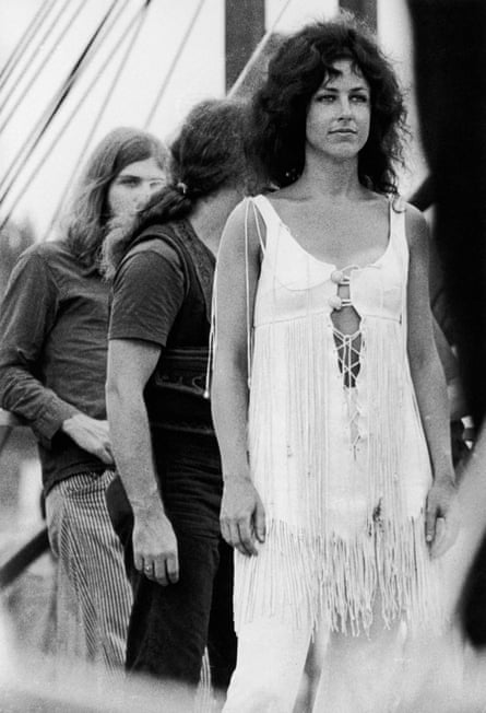 A black and white image of a woman with dark curly hair wearing a white fringed tunic dress and white trousers with a couple of men behind her