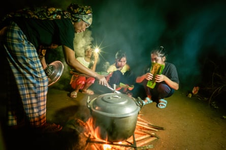 A woman bends over a large pot on a trivet on an open fire, while children squat nearby, watching