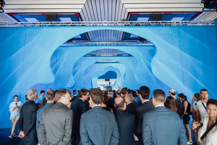 Men in suits can be seen at the back of large group of people looking away from the camera at a large blue artwork