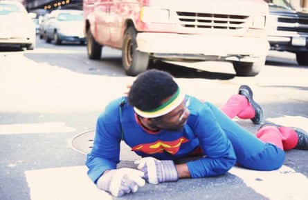 A man wearing a Superman costume crawling on the street, with cars behind him