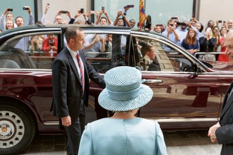 Queen Elizabeth II of England in 2014, back to the camera, walking towards a car on the other side of which are lots of spectators trying to take photographs of her