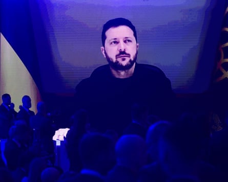 People in a darkened room watch Volodymyr Zelenskyy talking on a large screen