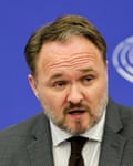he is in his late 40s with short, greying fair hair and beard; he has a round face and wears a grey jacket, white shirt and black tie as he speaks against a bright blue backdrop.