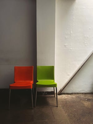 A green chair and an orange chair next to each other on a dark tiled floor, with a plain wall in the background