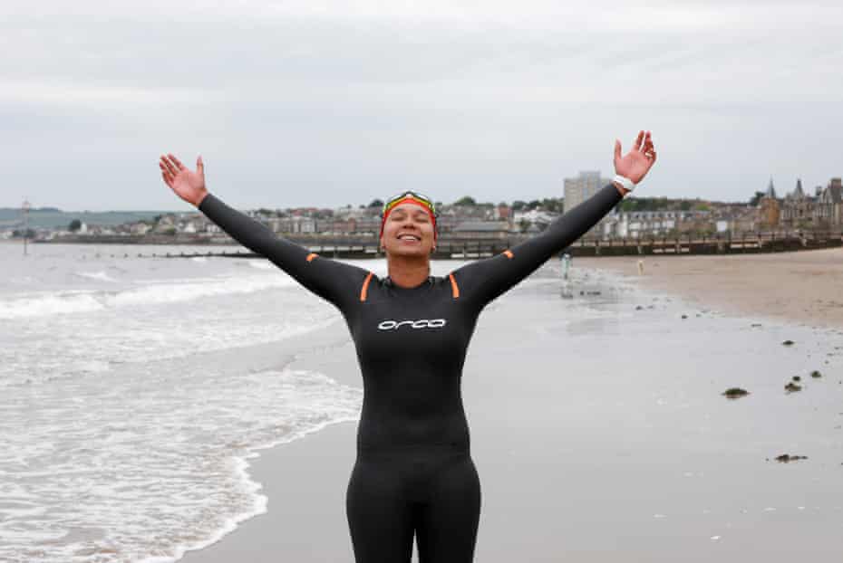 Alex Sehgal, wearing a black wetsuit, standing on the waterline with arms outstretched towards the grey sky