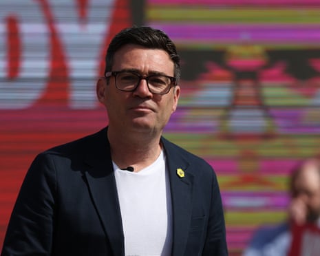 Andy Burnham in glasses and a dark blazer stands outside against colourful graffiti
