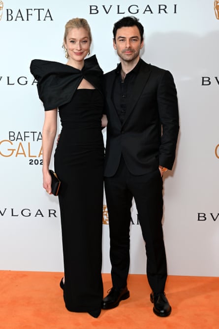 Caitlin Fitzgerald and Aidan Turner pose arm in arm on an orange carpet, both in formal black outfits