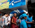 A Buddhist monk in red robes stands on a street with other Burmese people outside a shop marked Telenor