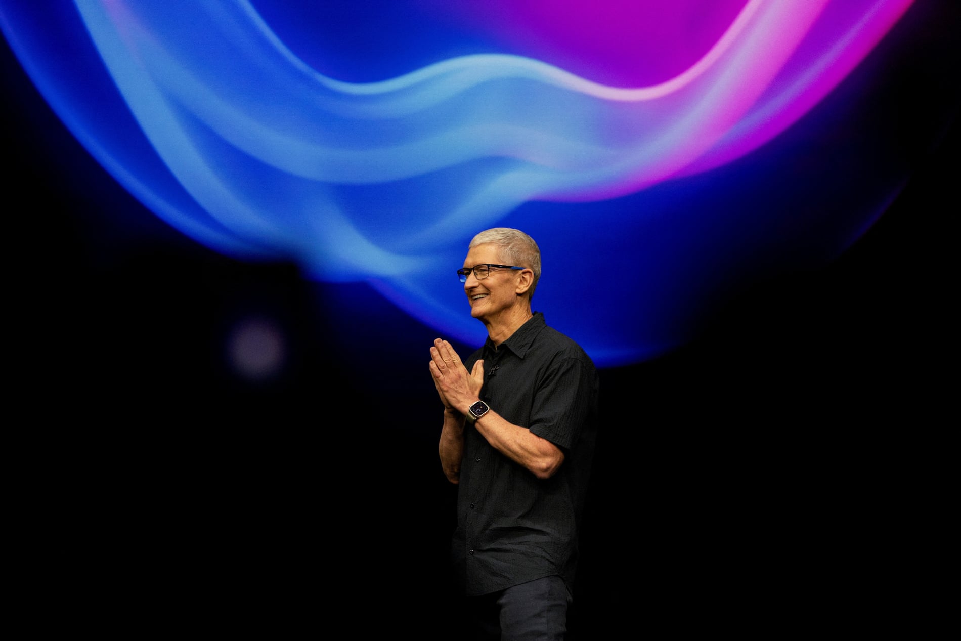 Tim Cook, CEO de Apple, aparece en el escenario durante un evento de lanzamiento de la compañía. Está vestido con una camisa negra y lleva puesto un Apple Watch. En el fondo, se observa una proyección de colores vibrantes en tonos azul y púrpura, creando un efecto de ondas luminosas. Cook sonríe mientras junta las manos, en un gesto de agradecimiento o bienvenida.