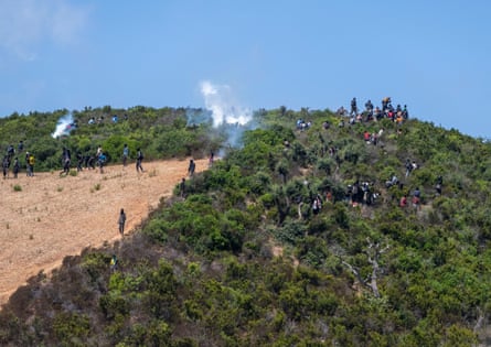 Wisps of white smoke rise from a hillside, in which dozens of people can be seen among plants and scrubland.