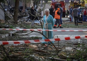A woman in a dressing gown stands among debris strewn in the street with police tape in foreground