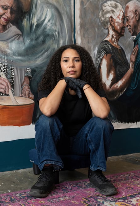 A young Black or mixed-race woman with long curly black hair squats in jeans and a black T-shirt in front of paintings