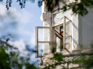 Chișinău, Moldova. A person in blue prison guard uniform looks out of a window on the corner of a white building with blurred branches of trees framing the top and bottom of the photograph