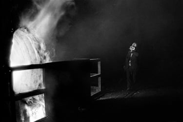 A steelworker in a protective mask stands near a blazing furnace amid smoke