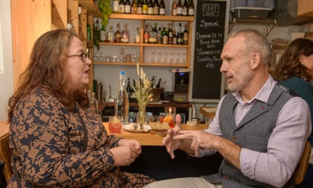 A woman with long red wavy hair, glasses and red lipstick, wearing a brown and black patterned dress, and a man with short grey hair, wearing a lilac shirt, a grey herringbone waistcoat and beige chinos, sitting at a restaurant table talking