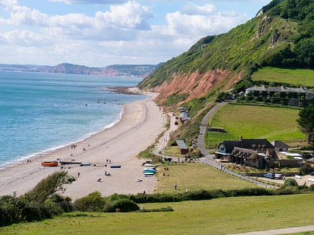 A beach with boats on the sand, beach huts, some buildings and grassy cliffs
