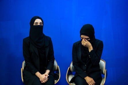 Two women sit in chairs wearing black hijabs and face coverings, against a blue background