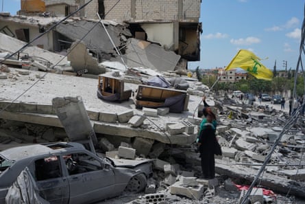 A woman handles a tattered yellow and green Hezbollah flag on the wreckage of her destroyed house. A wrecked car lies under rubble and slabs of concrete.