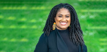 Bell Ribeiro-Addy: she is in her early 40s with long hair worn in narrow braids over her shoulders. She wears a black polo neck jumper and jacket plus large, colourful orange, green and yellow earrings, and stands against a bright green background with a big smile.