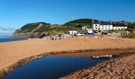 White buildings sit behind a golden pebble beach with green hills and sea in the distance.