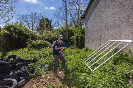 A man stands in undergrowth next to a house wall holding a fishing rod on a sunny day