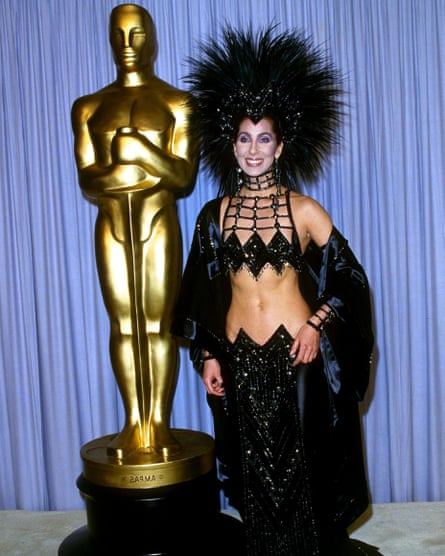 Cher standing next to a large gold Oscar statuette, wearing a dramatic black two-piece evening outfit with a beaded bra top, long embellished skirt and open robe and a striking spiked feathered headdress and bold choker-style jewellery.