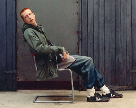 A male model sits on a chair in a green coat, jeans and black Veja trainers with a white V