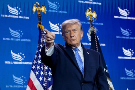 Trump in blue suit points his finger on a stage with two US flags and a blue background.