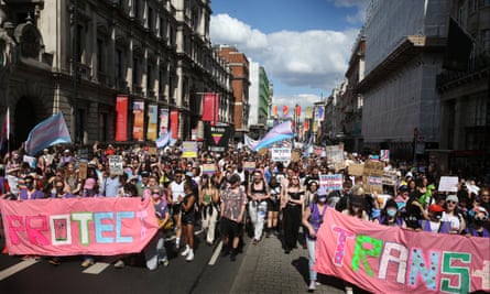 A large parade with people carrying colourful banners, with a large pink one in front saying ‘Protect trans …’
