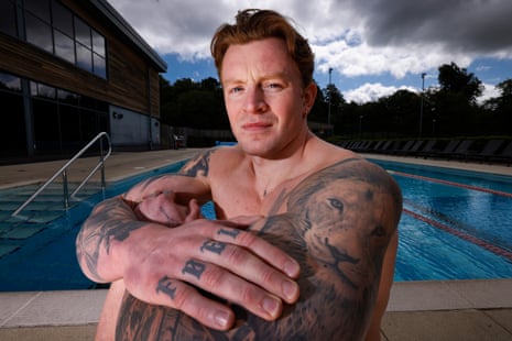 The British swimmer Adam Ramsay-Peaty, a white man with full sleeve tattoos on both arms and on his left hand, sits on the edge of a swimming pool with his knees up and arms crossed looking at the camera.