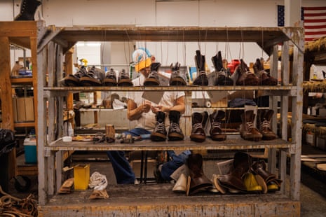 A man in a baseball cap, seen through an open shelving unit stacked with boots in various states of construction.
