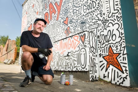 A man kneels in front of a graffiti-style mural with spray cans at his feet
