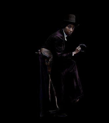 A woman wearing a 19th century style equestrian outfit against a black background.