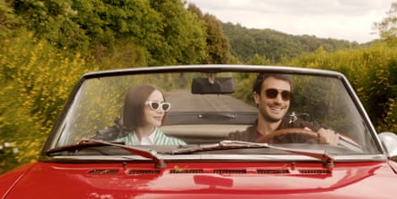 Emily and Marcello driving through countryside in a red convertible classic car. Emily is wearing white cat eye-style sunglasses and a green and white striped jacket