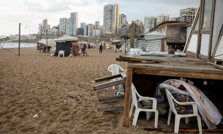 a makeshift encampment on a beach: tent-like structures have been erected as shelters on the sand, with white plastic chairs outside. Tall apartment buildings and towers are in the background.