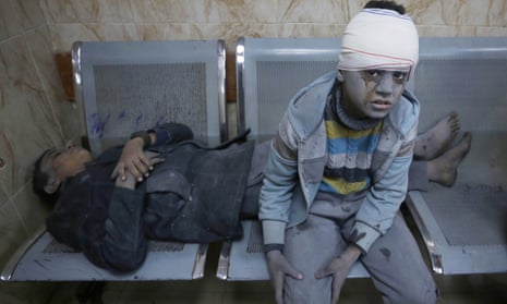 One boy lies on a bench behind another boy holding his leg, his head wrapped in a bandage and his face covered in grey dust