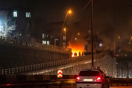 A fire burns on a street, with silhouettes of people who are gathered nearby.