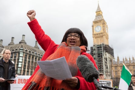 Abbott dressed in a red coat holds her fist in the air in the street. Big Ben is in the background.
