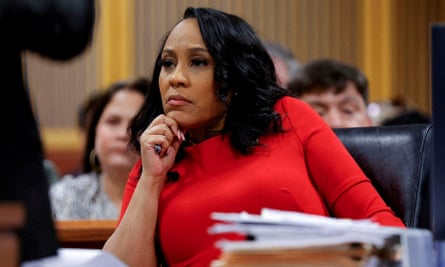 Middle-aged Black woman with bright red long-sleeved dress and shoulder length straight black hair rests chin on hand and appears to listen from behind large desk.