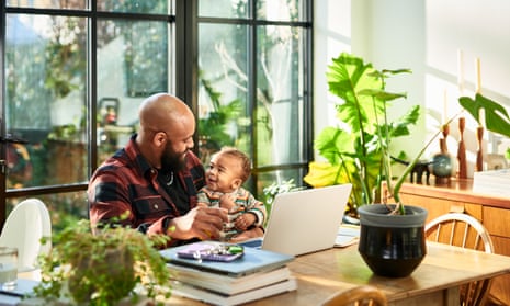 A young father with a baby sitting at a table in an airy kitchen in front of a laptop