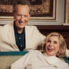 A woman smiling and leaning back on a sofa while a man smiles and stands behind her on the sofa