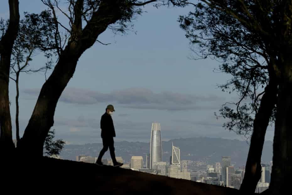 A person wearing a face mask walks atop Tank Hill in front of the San Francisco skyline in December.