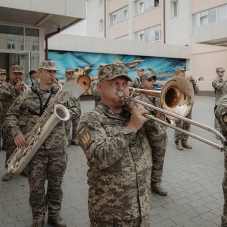 Military personnel in camouflage gear and caps play brass instruments including a trombone, trumpet and saxophone